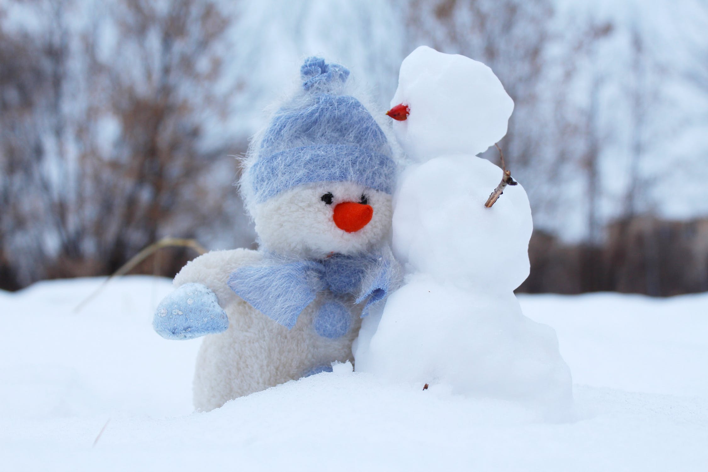 A picture of a fuzzy stuffed snowman toy with a blue hat and blue scarf next to a real snowball sized snowman