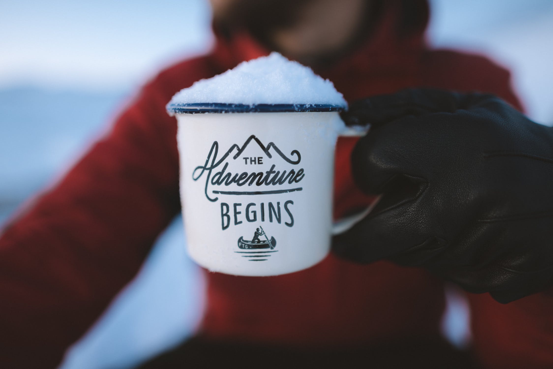 Tin cup filled with snowball with caption of The Adventure Begins held by man in red jacket