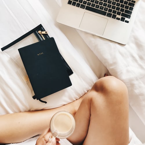 woman on bed with knees crossed, with a cup of coffee, laptop and planner ready for her morning routine