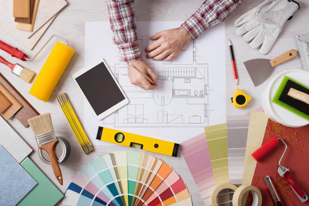 How To Increase Your Income by becoming an interior decorator. Decorator drawing on a house project with work tools, painting rollers and color swatches