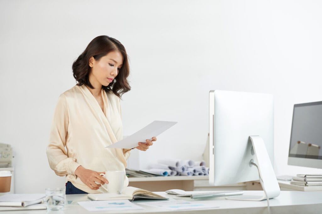 Online Proofreading Jobs Woman standing at computer desk while proofreading handful of papers